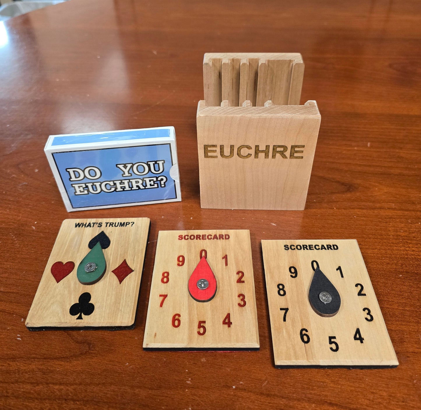 Euchre card game set on a wooden table, including a wooden deck box, a blue and white instruction card, and three scoring cards with heart-shaped numbers and playing card symbols.